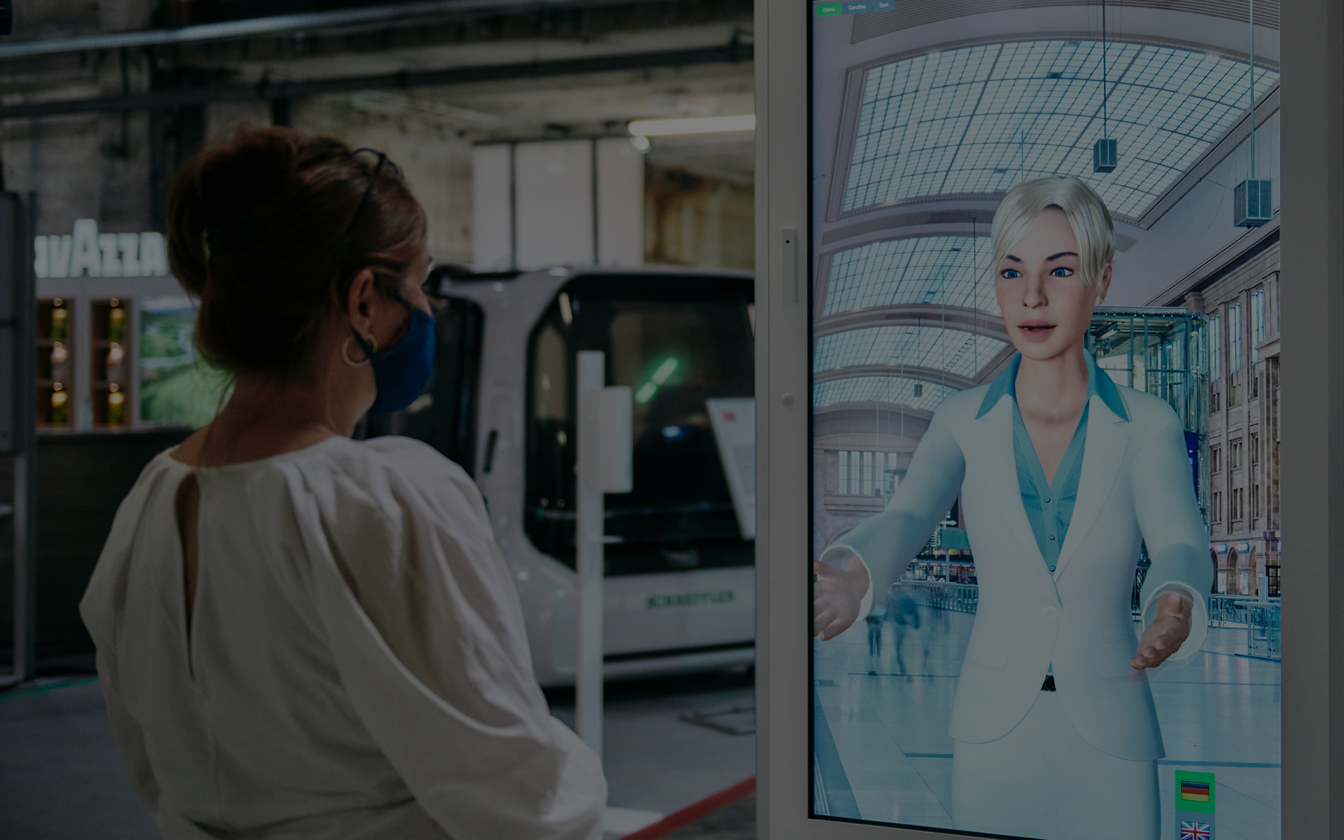 Frau interagiert am Bahnhof mit dem digitalen KI-Concierge DB SEMMI auf einem Bildschirm.