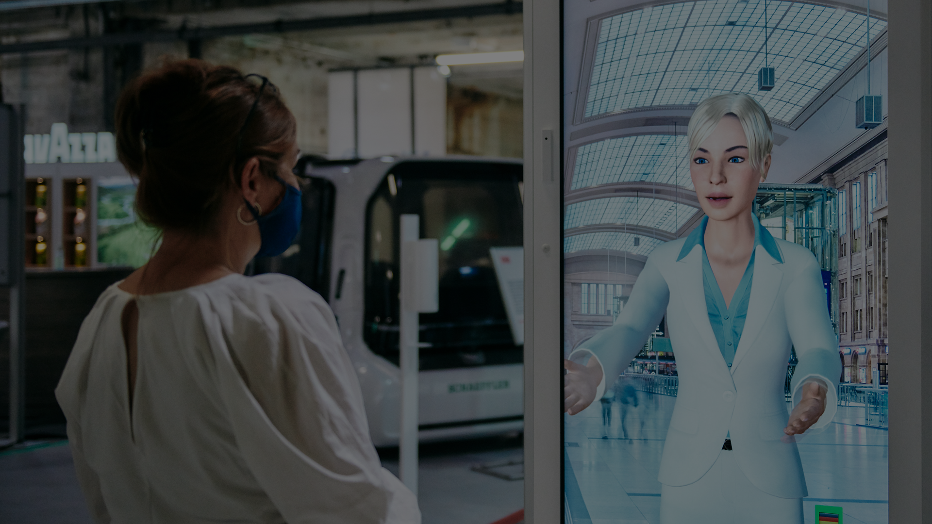 Frau interagiert am Bahnhof mit dem digitalen KI-Concierge DB SEMMI auf einem Bildschirm.