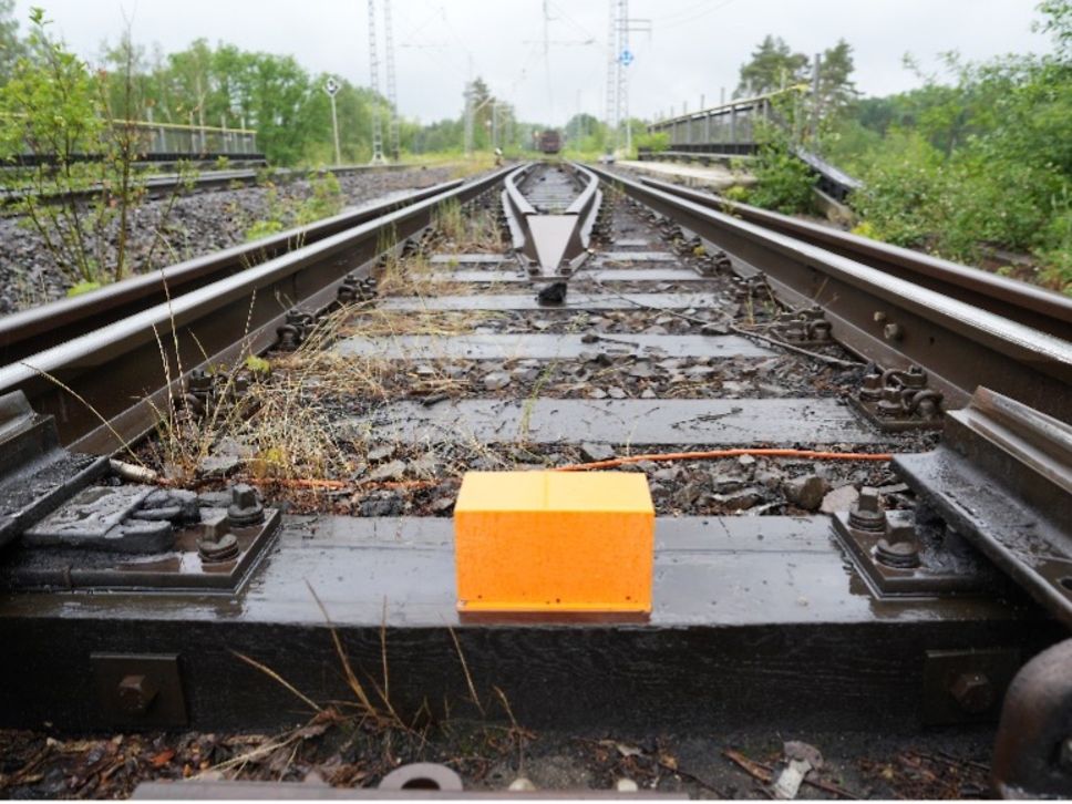 Orangefarbenes Messgerät steht auf einem Bahngleis zwischen Schotter und Vegetation.