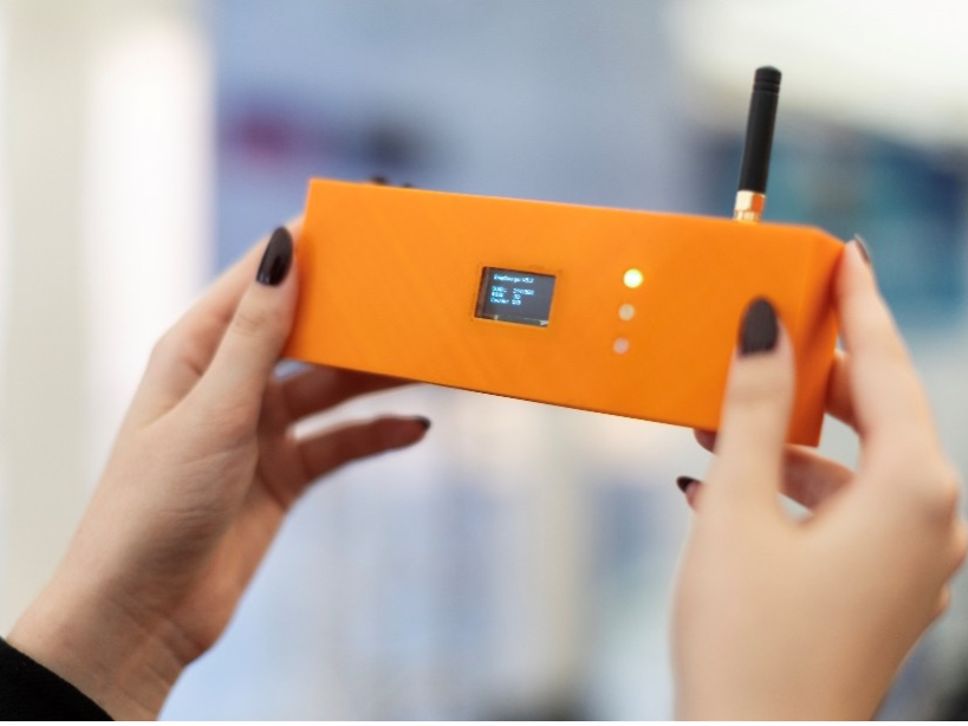 Hände halten ein orangefarbenes Messgerät mit Display und Antenne vor unscharfem Hintergrund.