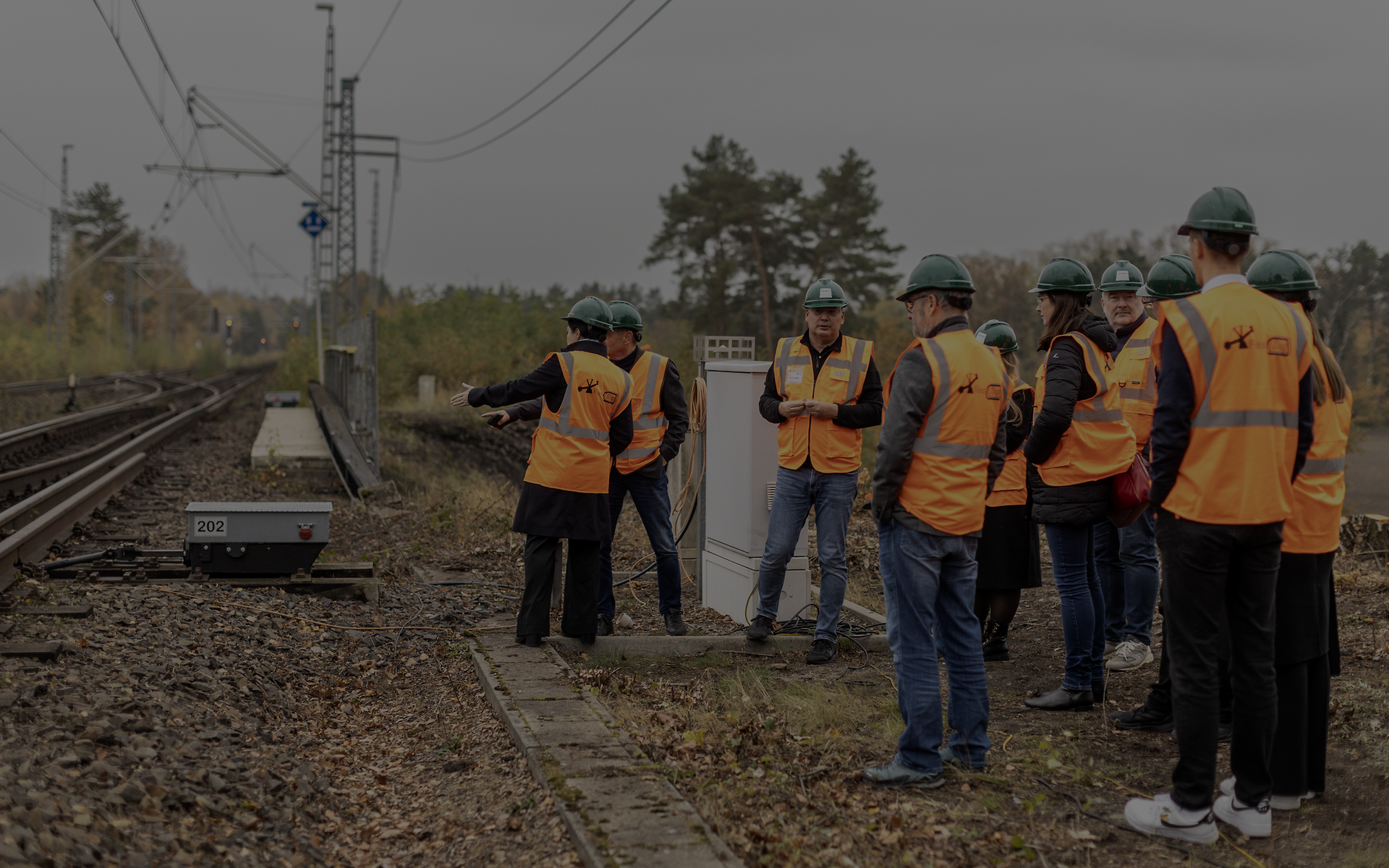 Gruppe mit Warnwesten und Schutzhelmen steht am Bahngleis und erhält eine Erklärung zur Bahntechnik.
